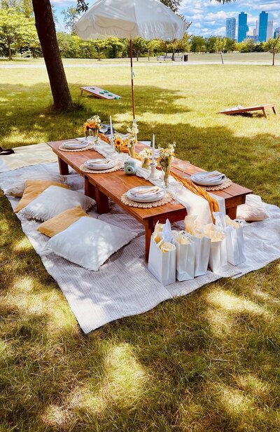 Outdoor picnic-style table with white cushions, floral centerpieces, and guest gift bags, styled by Yours Truly, a wedding planner and designer in New Jersey.