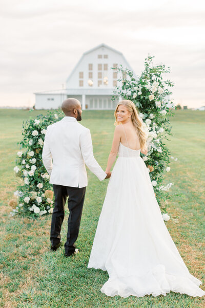 Stunning bride and groom walk towards luxe white barn wedding venue through floral arch on their wedding day at Providence Vineyard