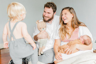 Dad and mom holding newborn  smile at toddler brother while sitting on the ground in Massachusetts Newborn Photography Session