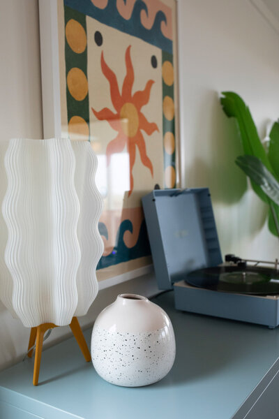 A stylish corner from a short term rental renovation featuring a blue record player, wavy white lamp, speckled ceramic vase, and a vibrant sun-themed art print on the wall.