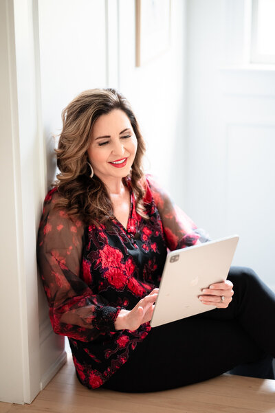 A woman with wavy hair wearing a red and black floral blouse sits on the floor leaning against a wall, smiling while using a tablet.