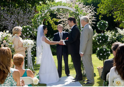 Conceive an idyllic setting with lush landscaping a joyous wedding couple, appreciative guests listening attentively to an officiant with crystal clear sound emanating from a professional sound system.