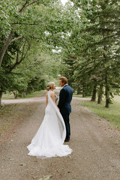 A bride and groom look at each other while standing in the middle of a tree-lined road in Calgary, Alberta