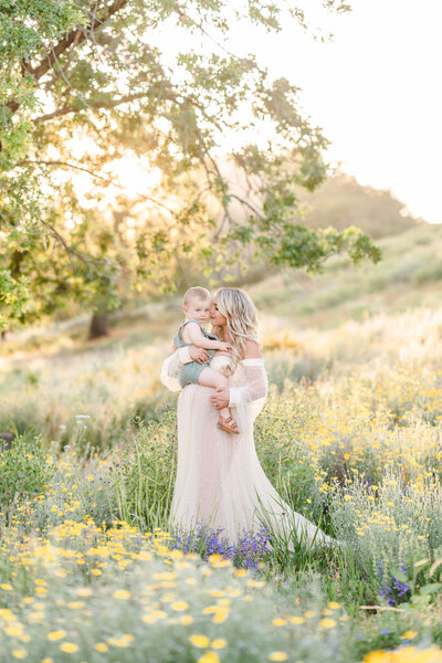 An expecting mother stands in a field of poppy flowers while holding her toddler child and gently placing her nose to his cheek photographed by Bay area photographer, Light Livin Photography.