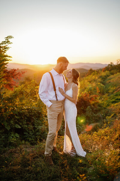 sunset elopement photos with couple standing on an outlook in the Foothills while the sun sets behind the Smoky Mountains as the couple embraces and smiles at one another as they elope to Gatlinburg 