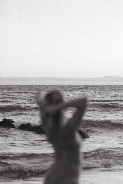 Artistic black-and-white photo of a blurred figure standing by the ocean, symbolizing stillness, self-connection, and emotional clarity through nature.