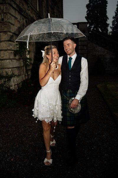 A couple walk linked together outside Haddo House at night. Newly married they are photographed under an umbrella as it rains. They smile and laugh and are dressed in wedding attire.