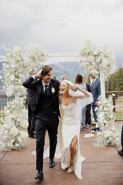 Newly weds in suit and wedding dress walk back down the aisle laughing. Backdrop is the vail mmountaions and a white floral covered cermony arch