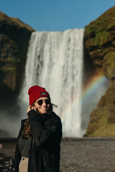 Female Destination Elopement Photographer in a red beanie and sunglasses smiling in front of Skógafoss waterfall in Iceland with a rainbow in the mist.
