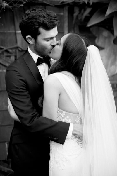 Black and white image of a man and a woman kissing. He's wearing a tuxedo, she's wearing a long white wedding dress with a veil.