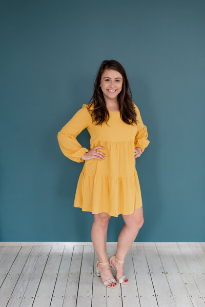 Bee, founder and owner of BusyBSocial, stares and smiles with her hands on her hips wearing a yellow dress in front of a blue background