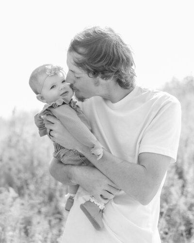 black and white photo of a father holding his 6 month old daughter giving her a kiss on her cheek as she's looking at the camera.  