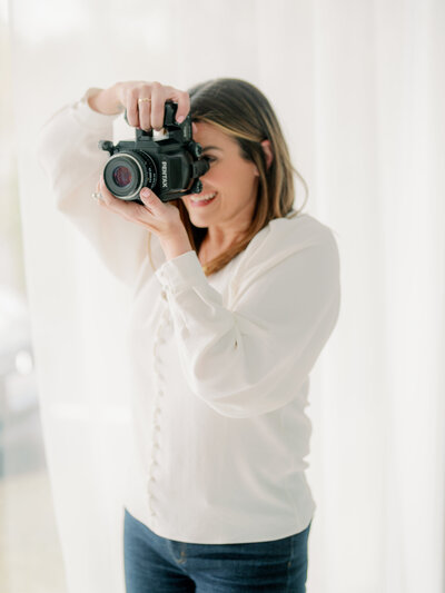 a woman in a white shirt holding a camera and photographing something and smiling