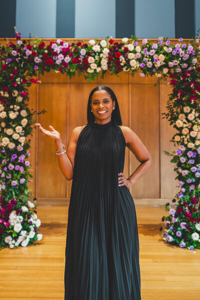 Black girl in black dress standing in front of  a  lush colorful floral  arch