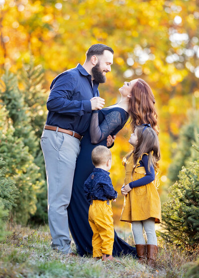 A husband and wife are dancing happily on a tree farm while their son and daughter watch them
