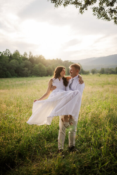 photographers in gatlinburg tn captures couple eloping to Gatlinburg in Cades Cove with groom carrying his bride through a meadow as her gown flows in the wind 