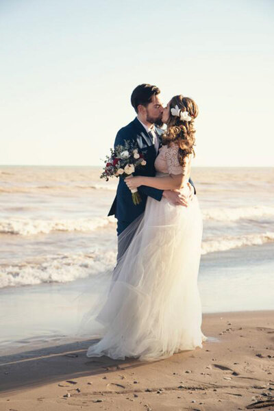 Married couple kissing each other while bride holding her floral bouquet on the seashore