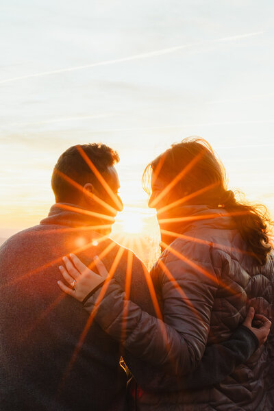 The sunset seen through two people who have their arms around each other and are smiling at each other 