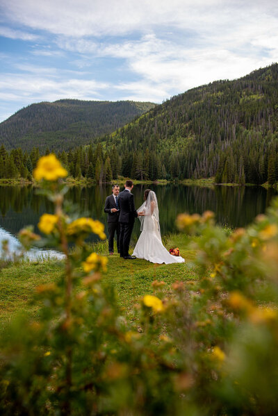 A bride and groom has a wedding ceremony  in front of a lake  surrounded by a yellow wildflowers and mountains in the background