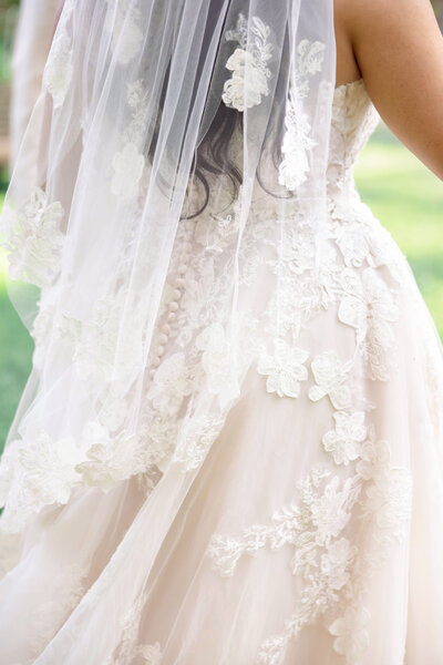 Close-up of a bride in a white wedding dress adorned with intricate lace details and floral patterns. The long veil drapes down the back, and soft curls of dark hair are visible. The background is a blurred green garden setting.