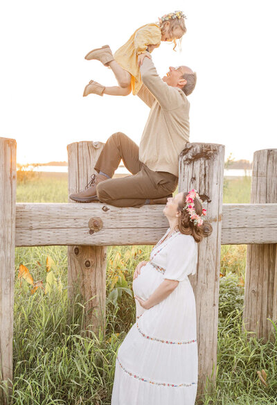 Dad sitting up on pier beam and holding toddler daughter in the air. Pregnant mom is standing below on the ground and looking up at both of them. Rooster Rock state park in the columbia river gorge family maternity photos.