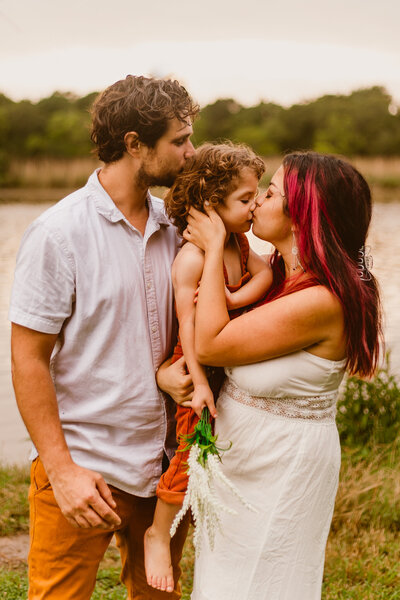 A family shares a sweet moment during a Mother’s Day picnic photo session at Bay Area Park in Texas. The mother kisses her young child while holding a bouquet, with a cozy white blanket spread out on the grass.
