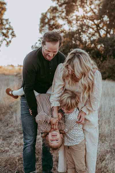 A family of four playing together while the dad holds his daughter upside down. 
