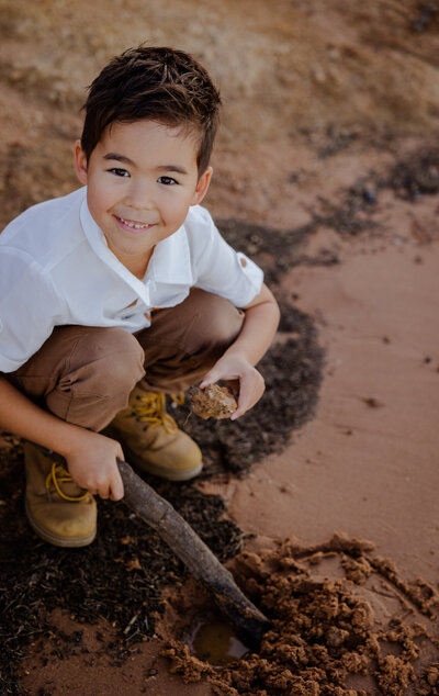 Smiling young boy playing in the sand in Thurgoona NSW, holding a stick and a rock while looking at the camera.