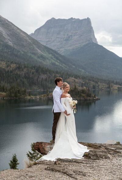 A couple stands on a cliff edge overlooking a calm mountain lake and dramatic peaks in Glacier National Park, the bride in an elegant off-the-shoulder gown and the groom in a white jacket, photographed by Sydney Breann Photography.