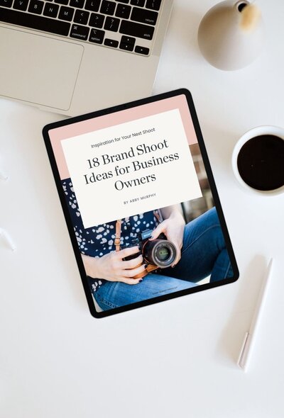 Laptop, tablet, cup of coffee, and a pen on a table with Abby Murphy's free download: 18 Brand Photoshoot Ideas for business owners on the tablet screen
