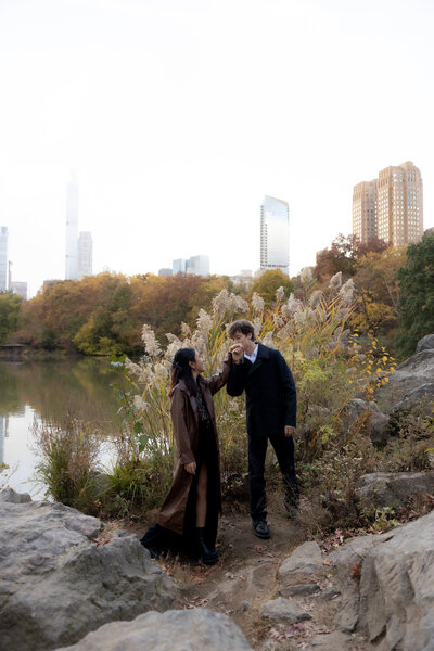 A couple standing together on a rocky ledge in Central Park with the New York City skyline in the background, captured during a fall couples session.