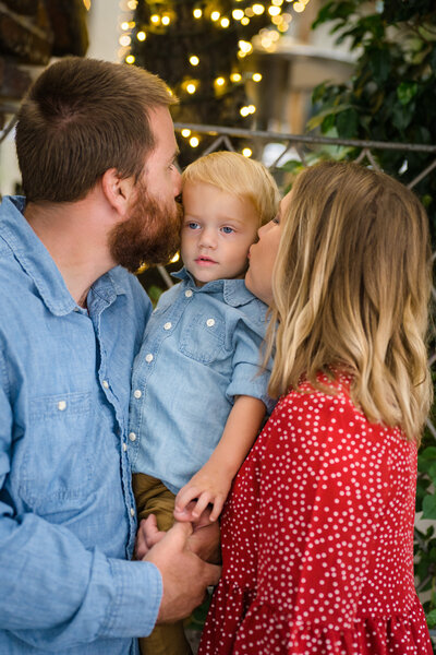 A mother and father hold their toddler son between them and kiss him on each cheek in front of twinkle lights. Photo by SAVI Photography - San Diego California Photographer