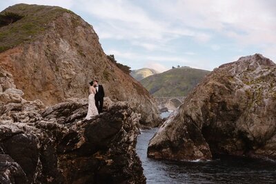 A couple in wedding attire stands on a rocky cliff in Big Sur with Bixby Bridge in the background.