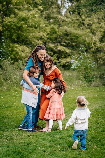 A joyful family of five, with the parents hugging their three young children outdoors on a grassy field, surrounded by lush green trees.