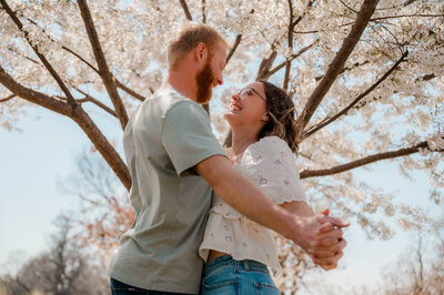 A couple holds hands and swings their arms in circles underneath a blooming tree.
