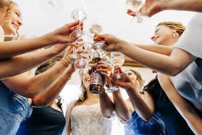 A group of women in dresses raise and clink glasses in a celebratory toast, with one woman holding a Coors Light beer bottle, viewed from below by an Eau Claire Wedding photographer.