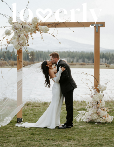 Bride and groom kissing with the bride’s veil blowing in the wind, set against a backdrop of mountains and a lake.