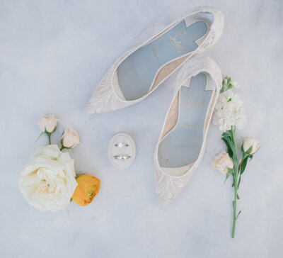 Photo of white lace wedding shoes on blue background with flowers around