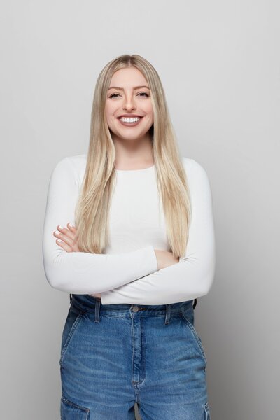 Portrait photo of Steph, a young white woman with blonde hair. She is smiling and has her arms folded. She is wearing jeans and a white shirt.