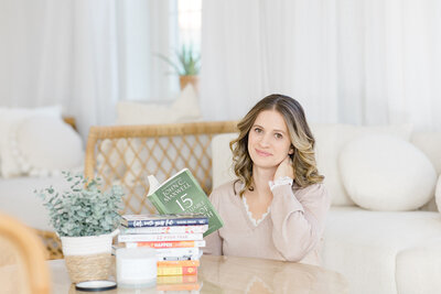 Woman sitting in white living room, smiling at the camera while looking through a pile of books.