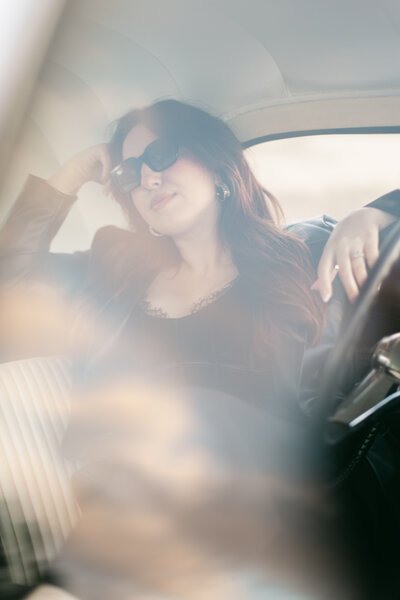 Moody, cinematic portrait of a woman wearing sunglasses inside a vintage white car. Soft reflections and dreamy light captured in Greenville, South Carolina by Dani Lacey Photographs.