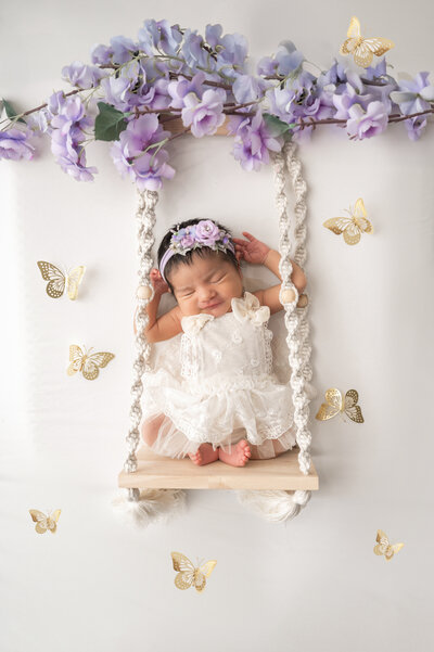 Baby laying on a white backdrop inside of a swing, surrounded by gold butterflies.