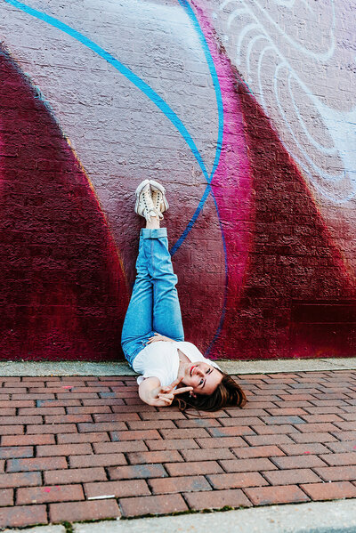 a senior girl laying on a colorful sidewalk with her  legs and feet up on the wall