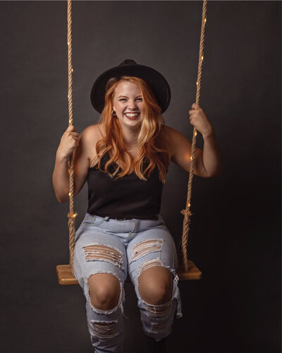 laughing orange-haired lady in a black top, black hat and ripped blue jeans sitting on a lit wooden swing in front of a dark background