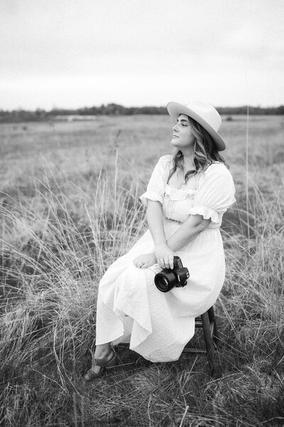 woman sitting in a field wearing a dress and hat while holding a camera