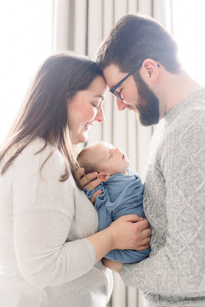 Man and woman forehead to forehead while looking down and their newborn baby wrapped in blue.