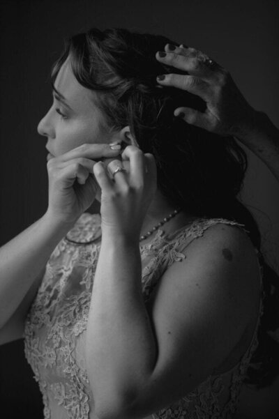 A bride stands at a window putting on her earrings with mother's hand in her hair