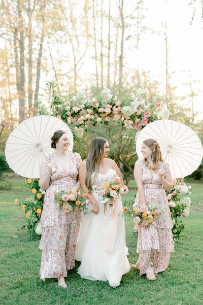 Bride with bridesmaids holding parasols and garden-style bouquets under floral arch.