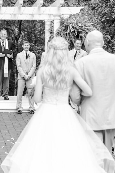 B&W image of a bride walks down the aisle to her groom while holding onto her fathers arm.
