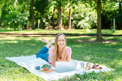 Emily Howell lies on a picnic blanket in a park and works on her computer, smiling